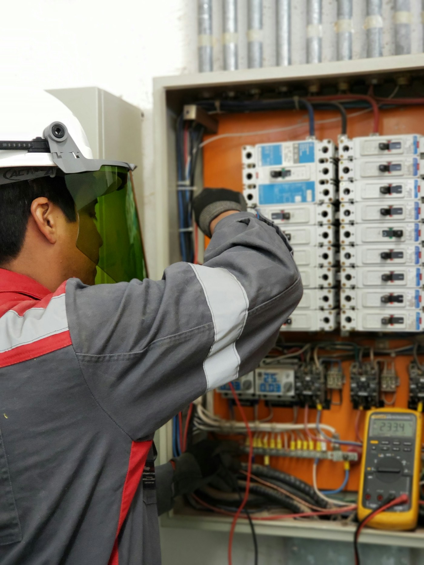 Electrician in a face shield and insulated gloves testing circuit breakers inside an electrical panel